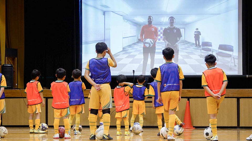 「KDDIみらい共創プログラム」の第一弾、「遠隔サッカー教室」の模様。京都サンガF.C.のマルコ・トゥーリオ選手が「空間自在コネクター」で子どもたちを“直接”指導